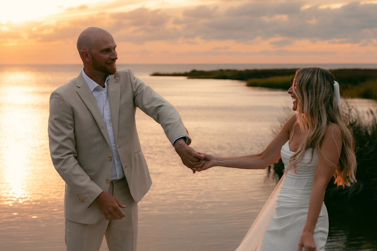 Newlyweds holding hands and laughing at the water's edge at sunset