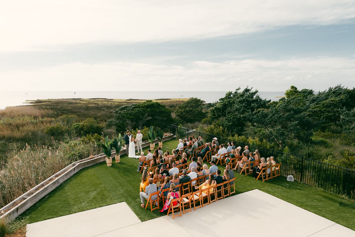 Aerial view of guests seated on a lawn for a ceremony beside the sound and marsh