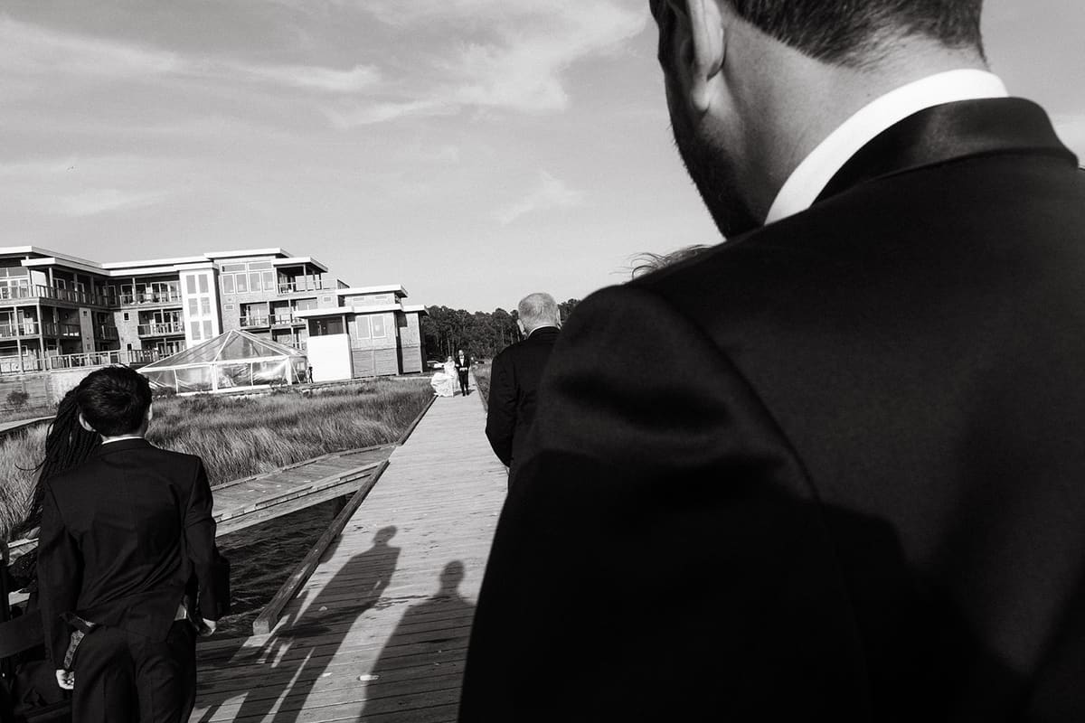 Black-and-white photo of the groom waiting on a boardwalk as the bride approaches